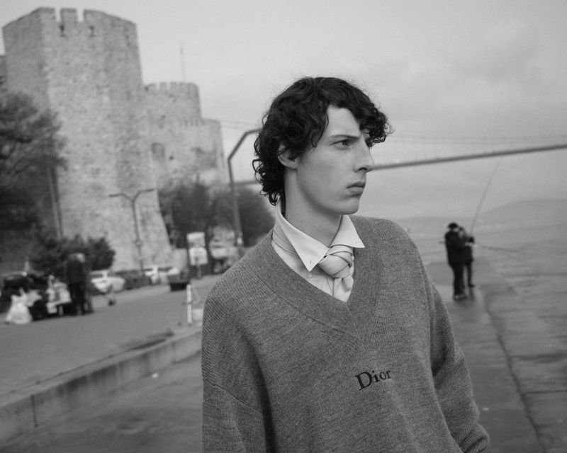 Monochrome fashion portrait of a male model wearing a grey V-neck sweater and a white collared shirt outdoors by a historic stone fortress