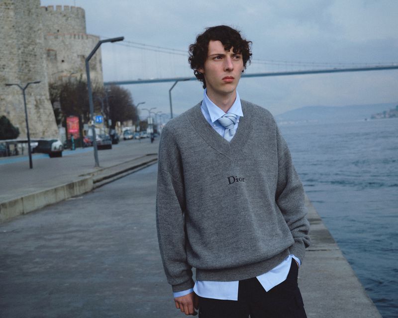 Model standing by a waterfront promenade wearing a grey v-neck sweater over a light blue shirt with a tie