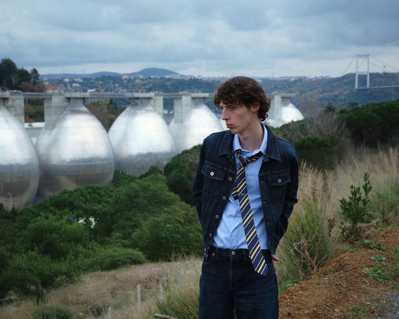 Young man in dark denim jacket and striped tie standing outdoors near large industrial domes and a bridge