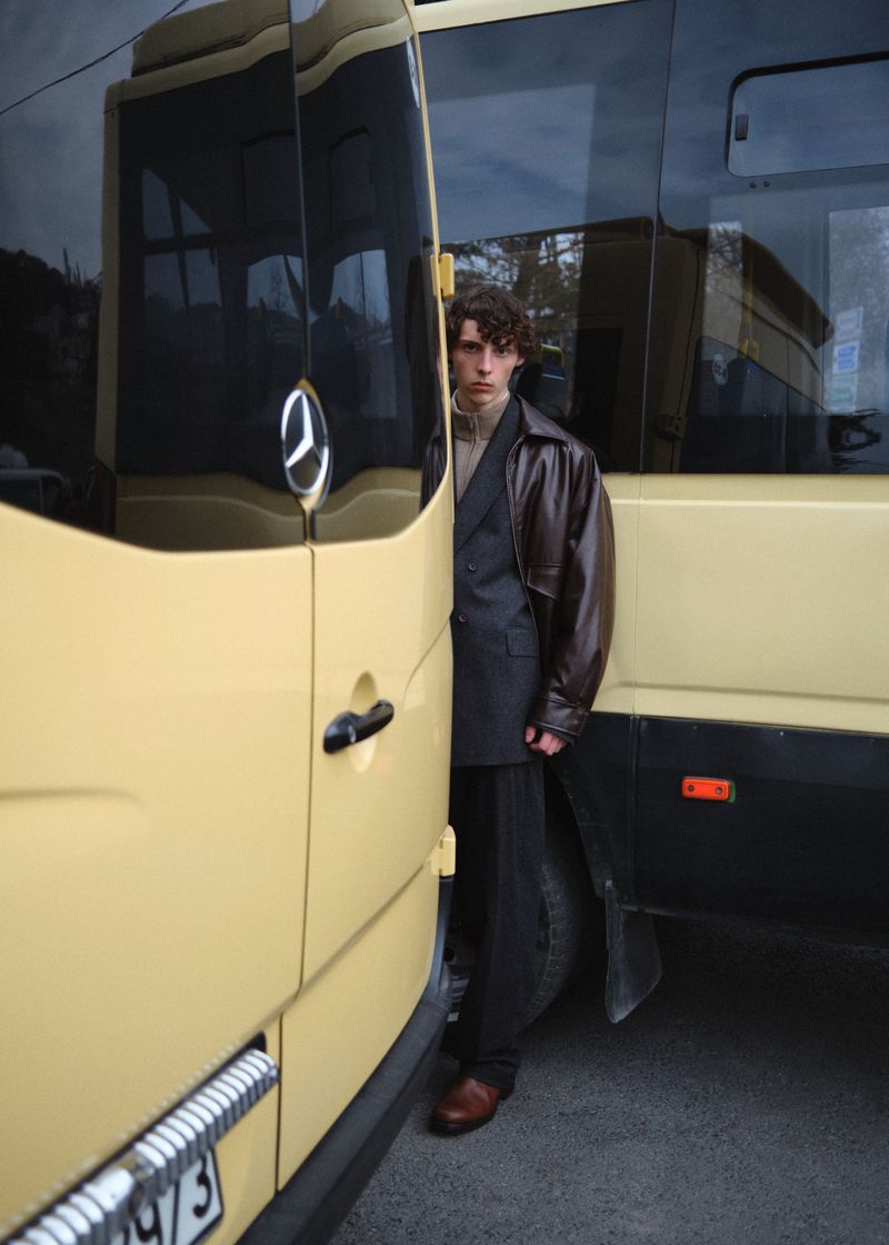 Model standing between two yellow vehicles wearing a dark leather jacket over a blazer and trousers.