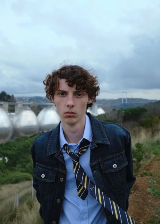 Young man with curly brown hair wearing a dark denim jacket, light blue shirt, and striped tie outdoors under a cloudy sky.