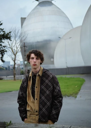Young man with curly hair wearing a plaid jacket and tan corduroy shirt standing outdoors before large spherical structures