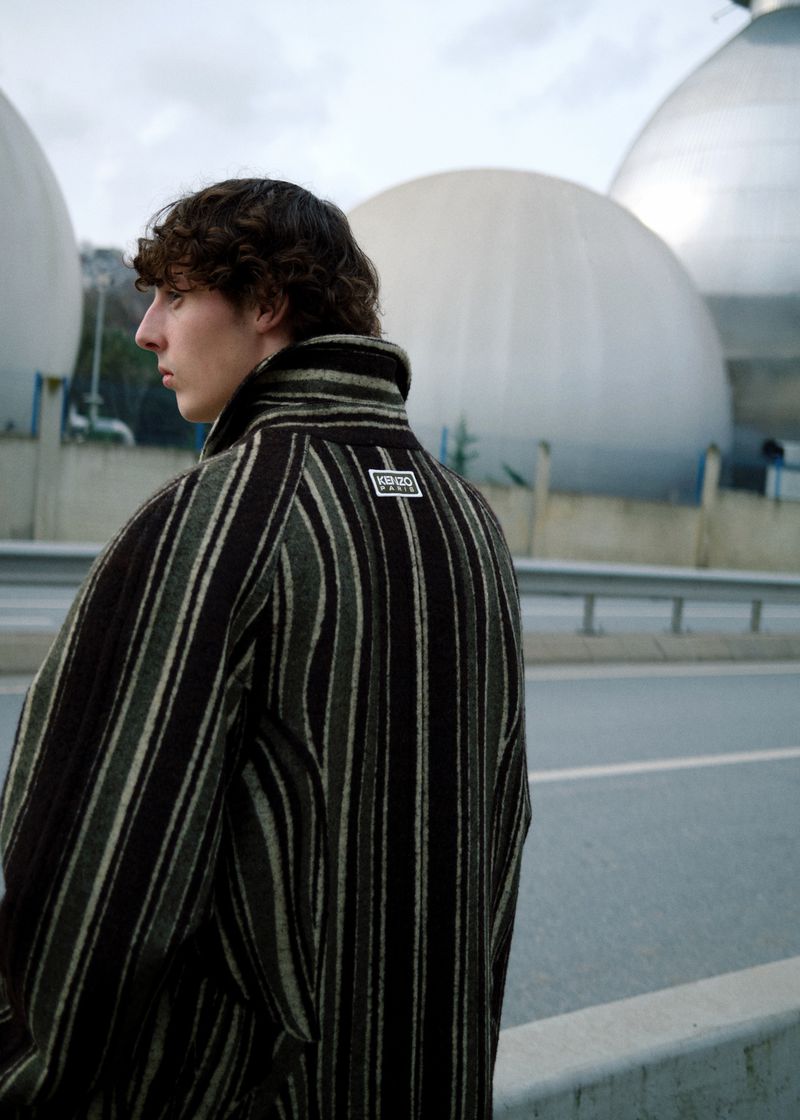 Young adult with curly brown hair wearing a striped wool coat stands on a road with concrete spheres in background under an overcast sky.