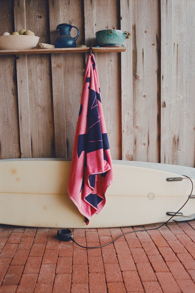 Pink and navy patterned beach towel hanging over a surfboard against a wooden wall and red brick ground