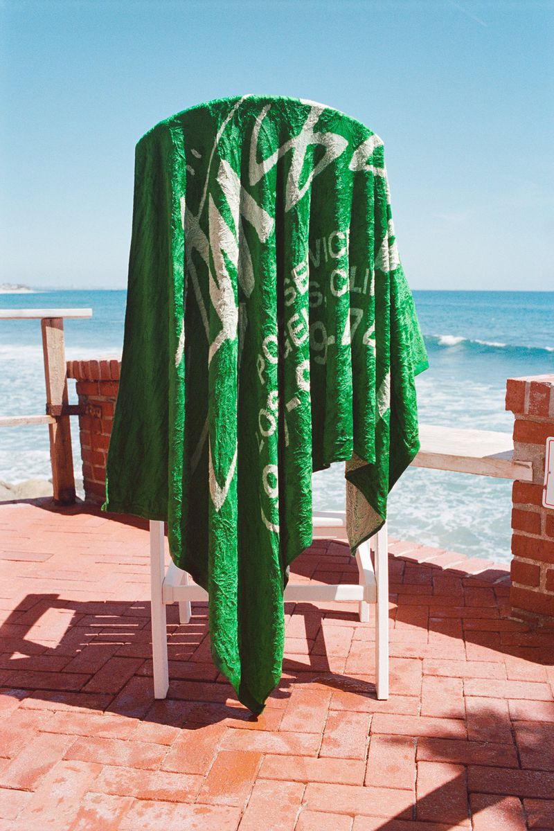 Green Stussy beach towel draped over a white chair on a brick patio overlooking the ocean under a clear sky.