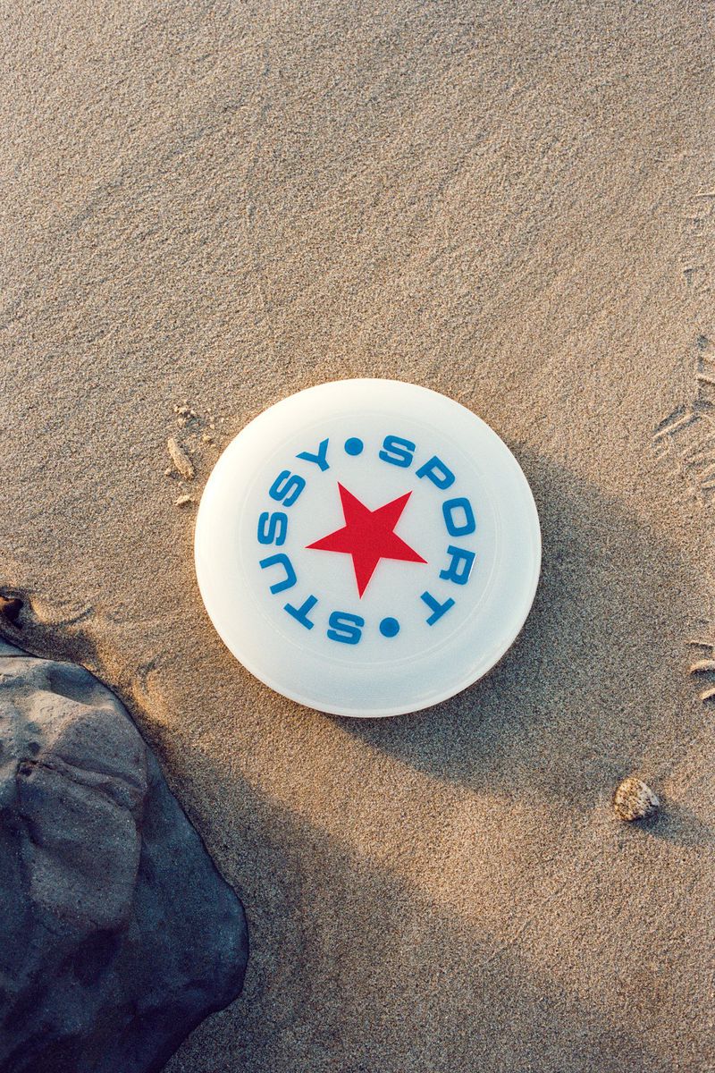 Overhead shot of a white Stussy Sport frisbee with a red star lying on a sandy beach next to a dark grey rock on a sunny day.