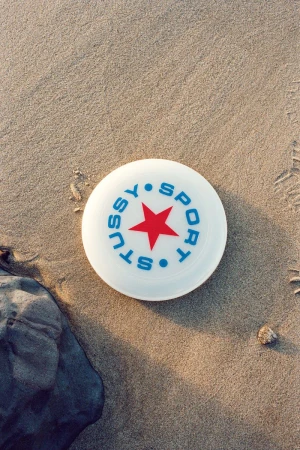Overhead shot of a white Stussy Sport frisbee with a red star lying on a sandy beach next to a dark grey rock on a sunny day.