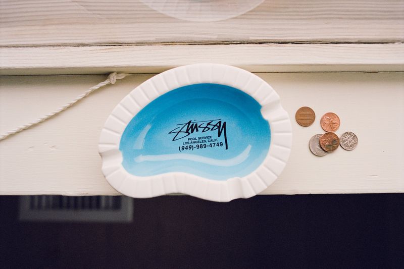 Ceramic swimming pool shaped ashtray with blue interior and text resting on a light wooden surface next to loose coins