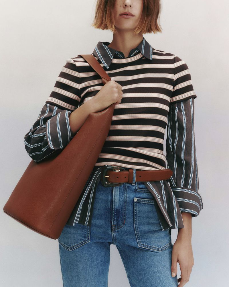 Model wearing striped top and denim jeans holding a large brown leather shoulder bag against a neutral studio backdrop.