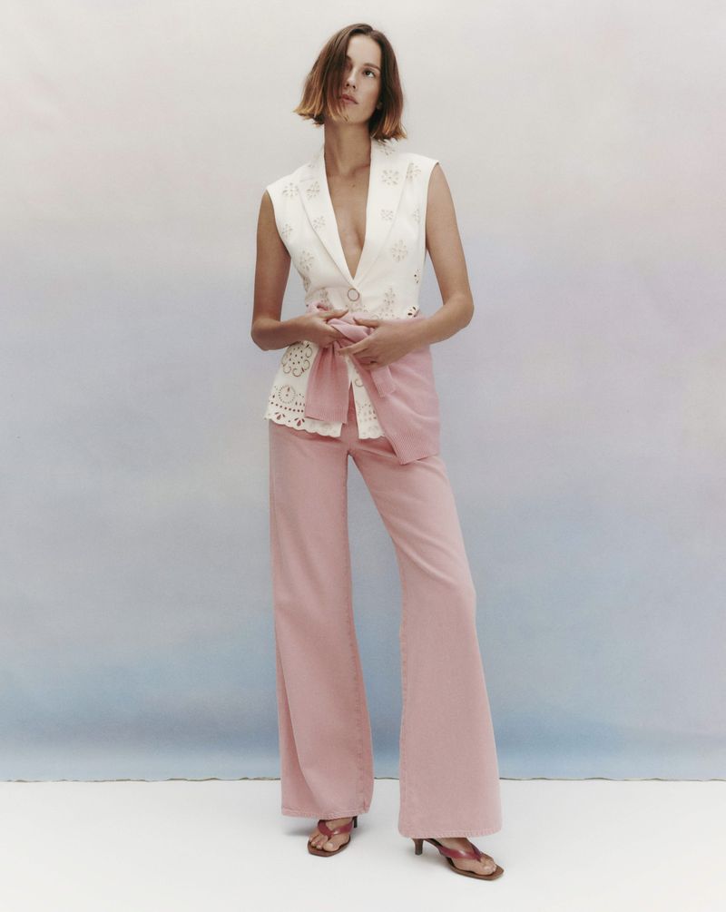 Model Mali Koopman in a white sleeveless patterned vest and pink trousers standing against a pale studio backdrop.