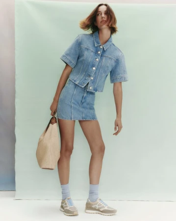 Model wearing a matching denim shirt and skirt holding a tan handbag against a light green background.