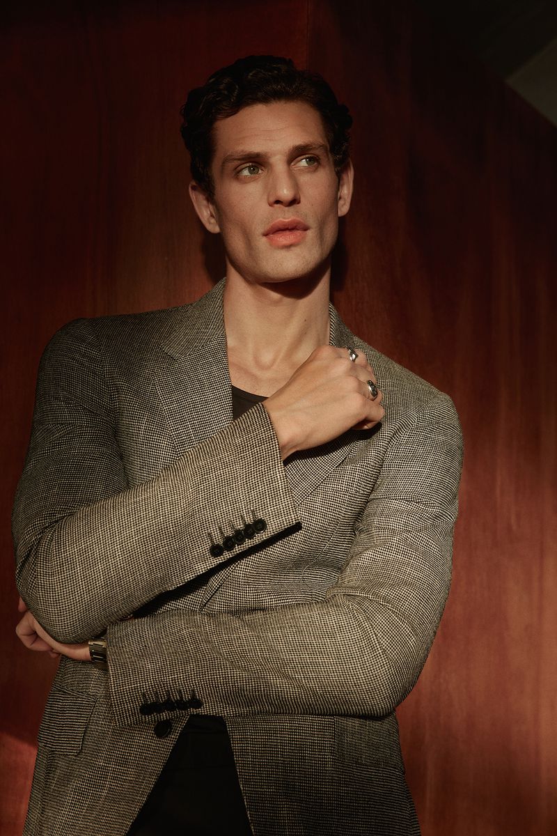 A man wearing a grey textured blazer posing against warm reddish-brown wood paneling for Calibre AW26 Lookbook.