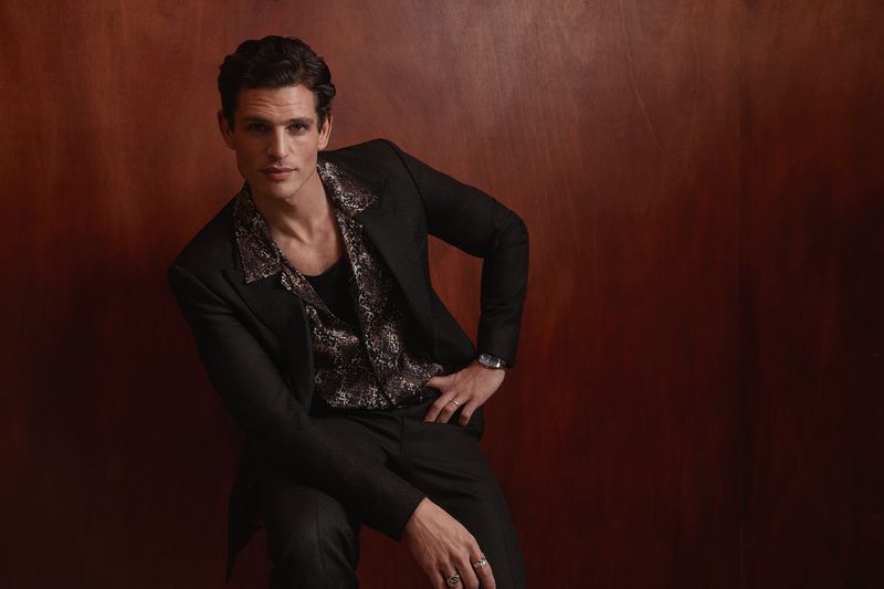 Portrait of a stylish adult male in a black suit and patterned shirt, posing indoors against a warm, reddish-brown background.