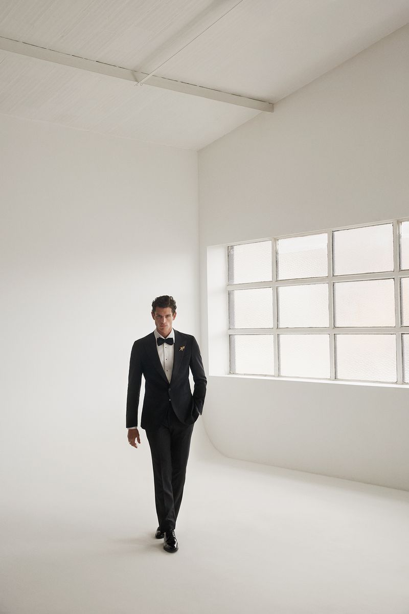 A man in a black tuxedo and bow tie walking across a white studio space with large windows in the background.