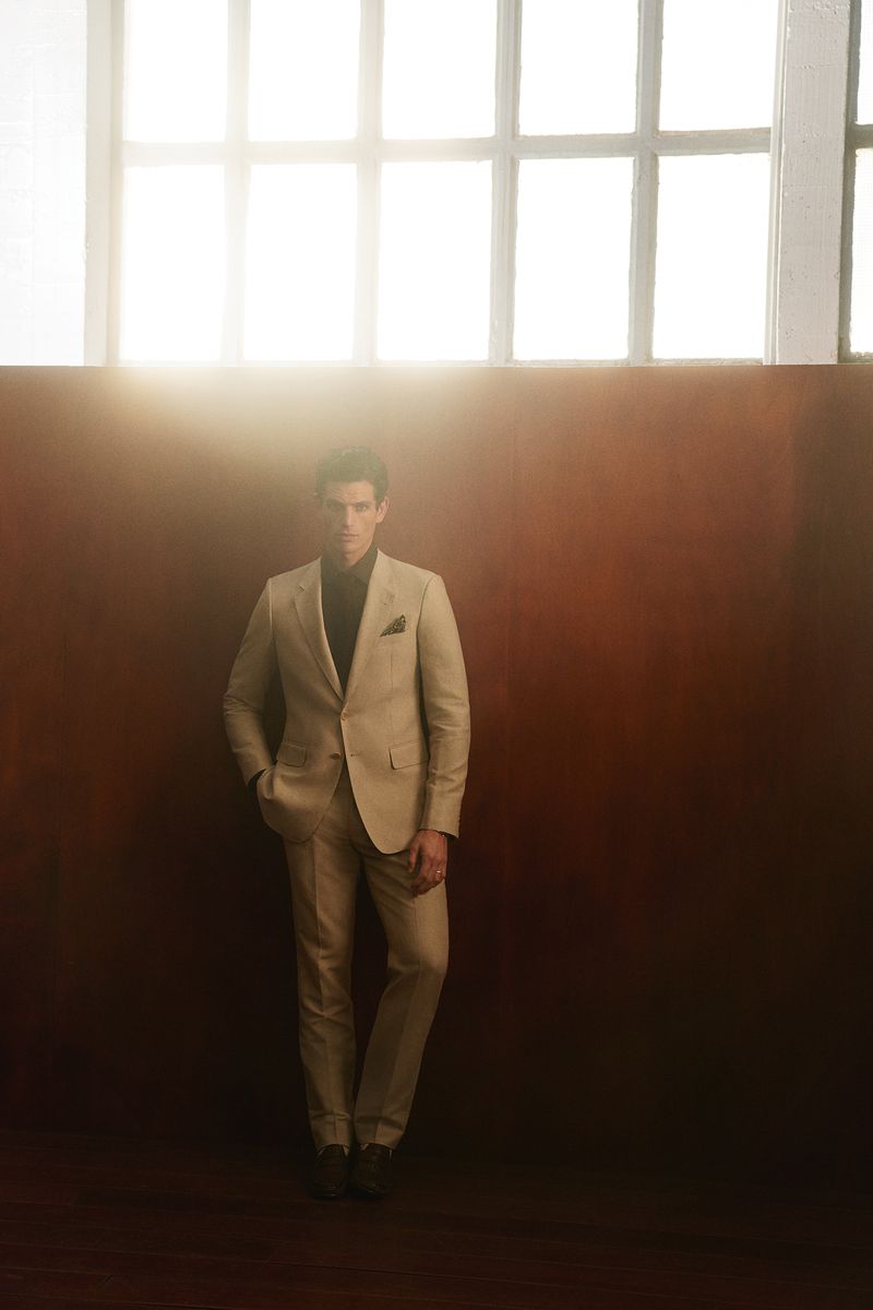 Man wearing a tailored beige suit standing against a dark wall beneath large industrial windows with bright backlight