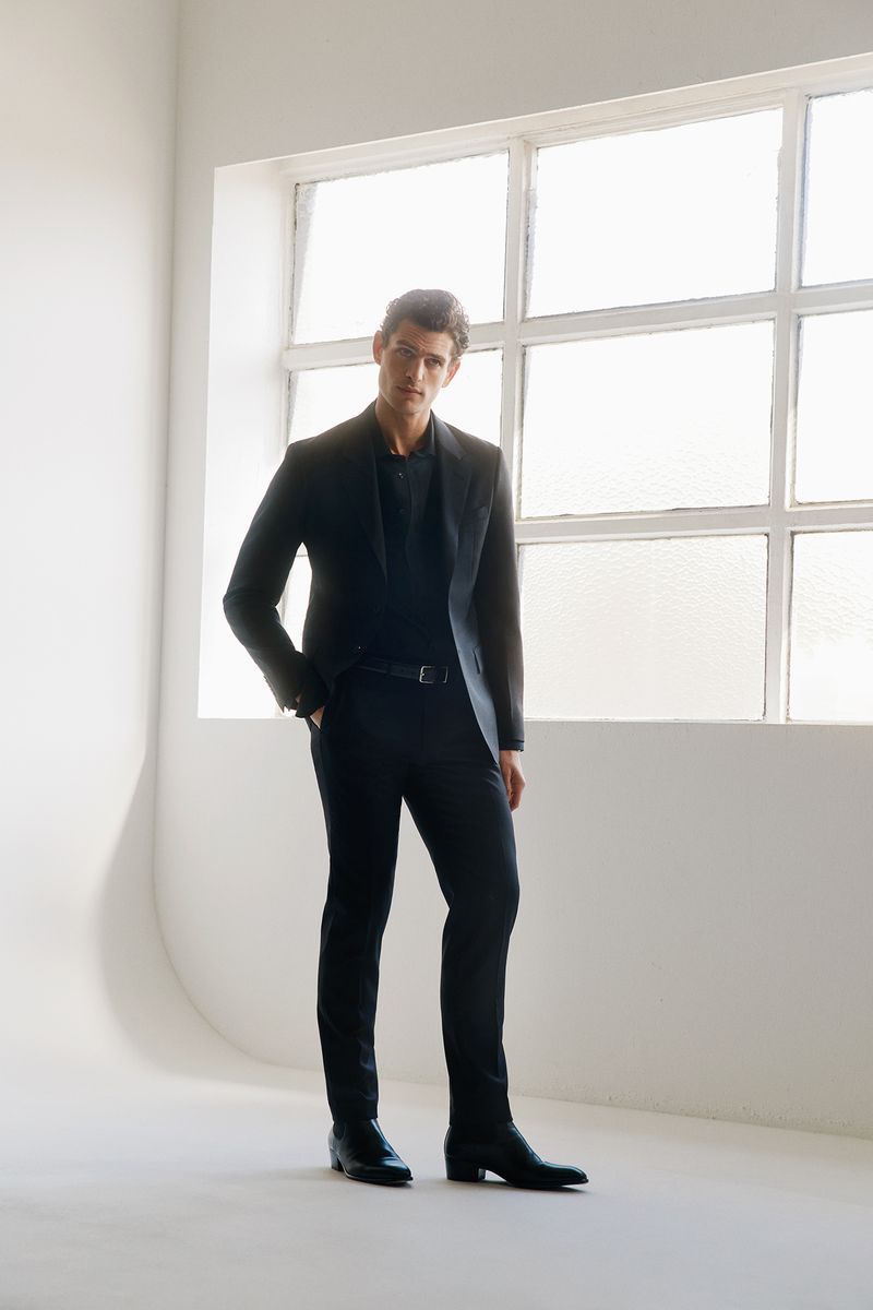 A male model standing in a dark suit against a white studio wall with a large gridded window.