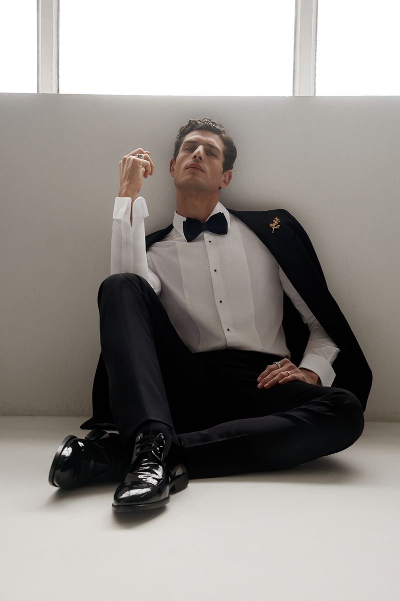 Male model seated on floor wearing white tuxedo shirt, black bow tie, black trousers and patent leather shoes.