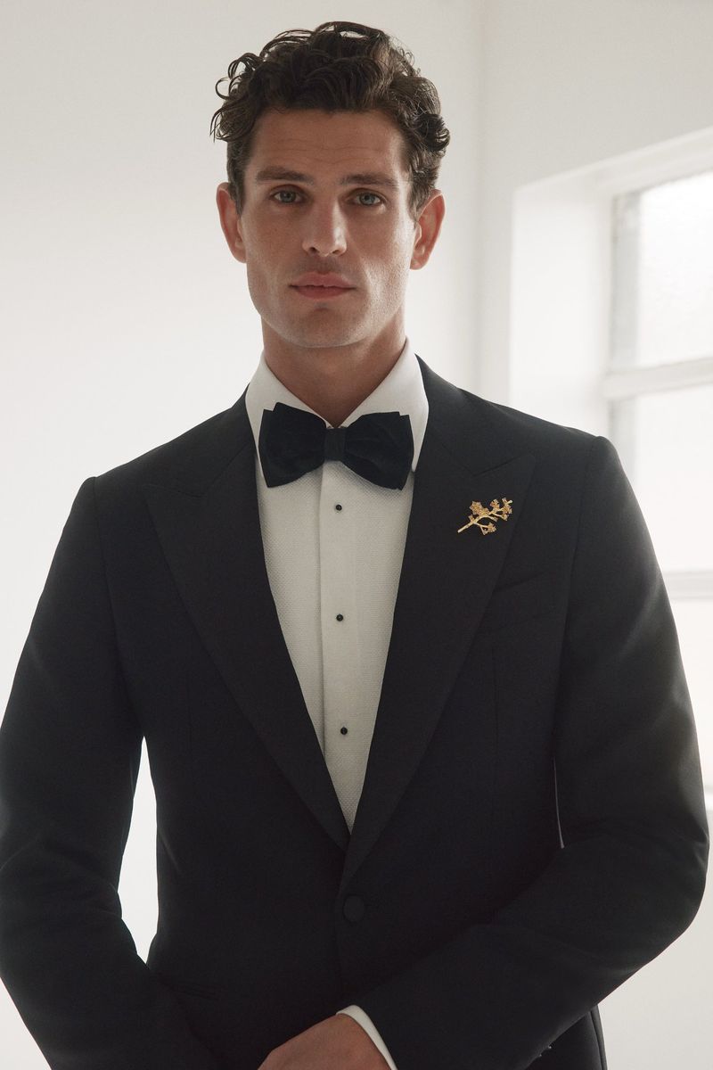 A man wearing a black tuxedo jacket, white shirt, and black bow tie with a gold lapel pin against a bright window light.