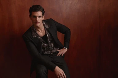 Male model wearing a black suit jacket and patterned shirt posing against a rich wood texture background.