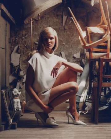 Woman in white dress crouching indoors surrounded by decaying walls and stacked wooden chairs, showcasing fashion in a distressed environment.
