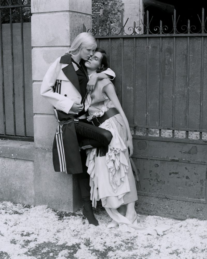 Black and white shot of two female fashion models in embrace in front of a stone wall and wrought iron fence in an outdoor setting.