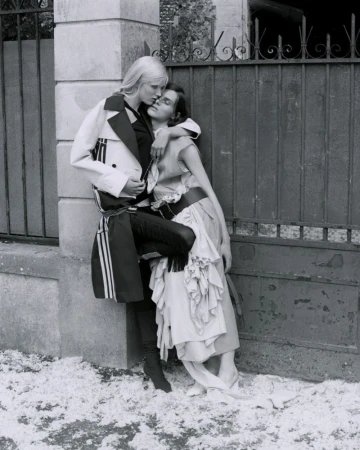 Black and white shot of two female fashion models in embrace in front of a stone wall and wrought iron fence in an outdoor setting.