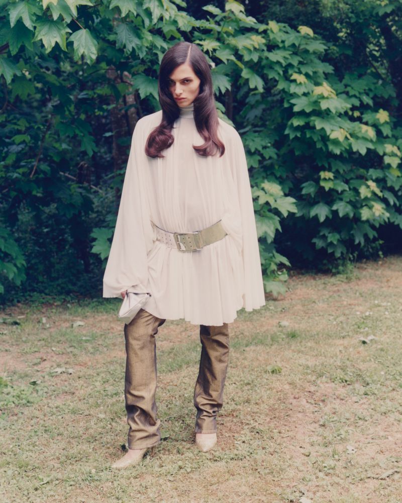 Woman in a flowing white tunic, gold pants, and matching pointed shoes poses outside in a green, leafy garden setting with natural light.
