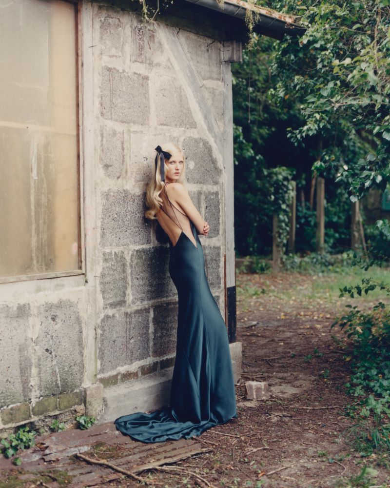 Woman in long teal dress poses against a block building with trees in the background, showcasing fashion in a natural outdoor setting.