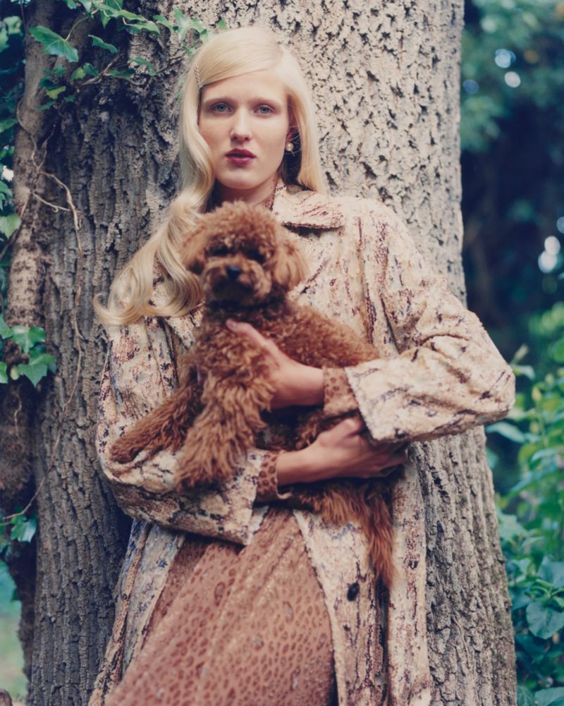 Adult woman in patterned coat poses with a brown poodle in front of a tree in a wooded outdoor setting on a sunny day.