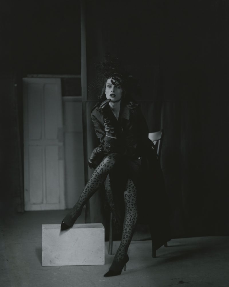 Monochrome portrait of a woman in a dark jacket, animal-print tights, and heels, posed in a dimly lit photo studio with sparse props.