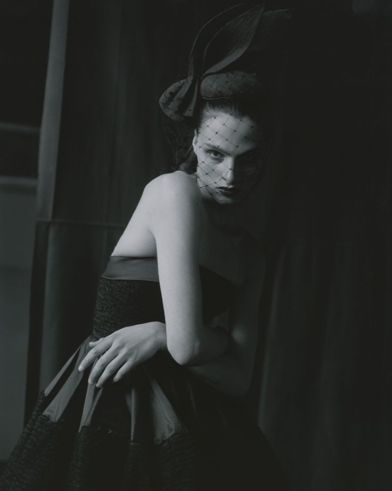 Elegant woman poses in strapless dress and net fascinator hat in a low-key, dark studio environment, creating a moody, fashion-forward image.