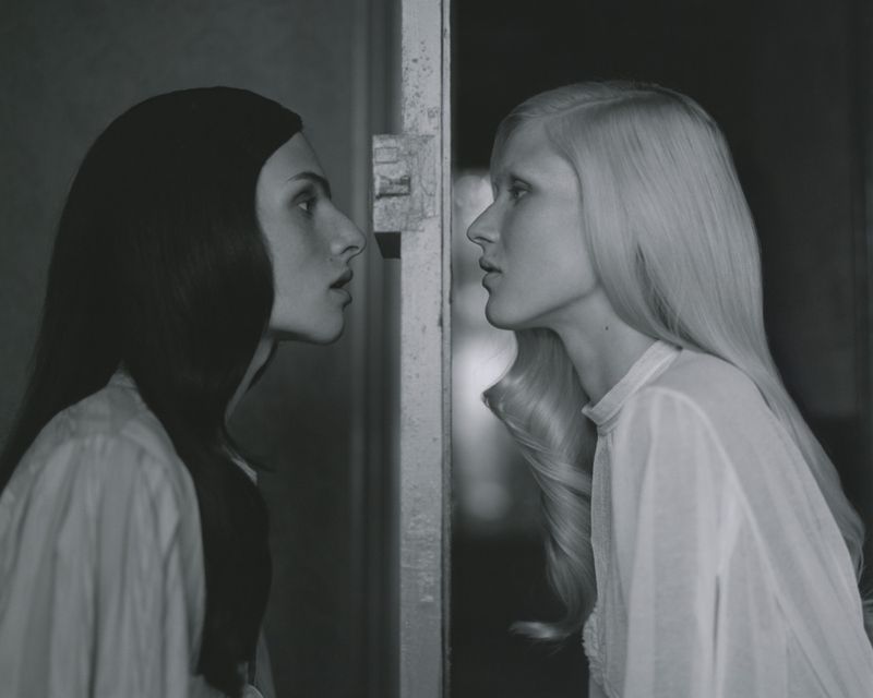Monochrome portrait of two fair-skinned women facing each other, one with dark hair and one with light hair, in a dimly lit indoor space.