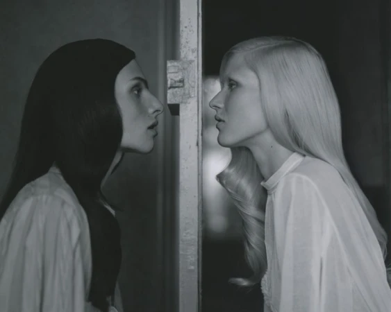 Monochrome portrait of two fair-skinned women facing each other, one with dark hair and one with light hair, in a dimly lit indoor space.
