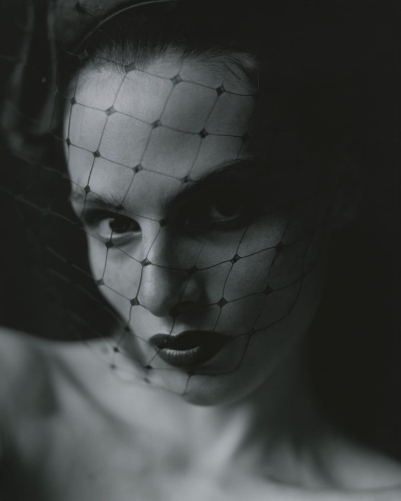 Monochrome portrait of a woman with a net veil over her face, highlighting her eyes and lips against a dark, blurred background.