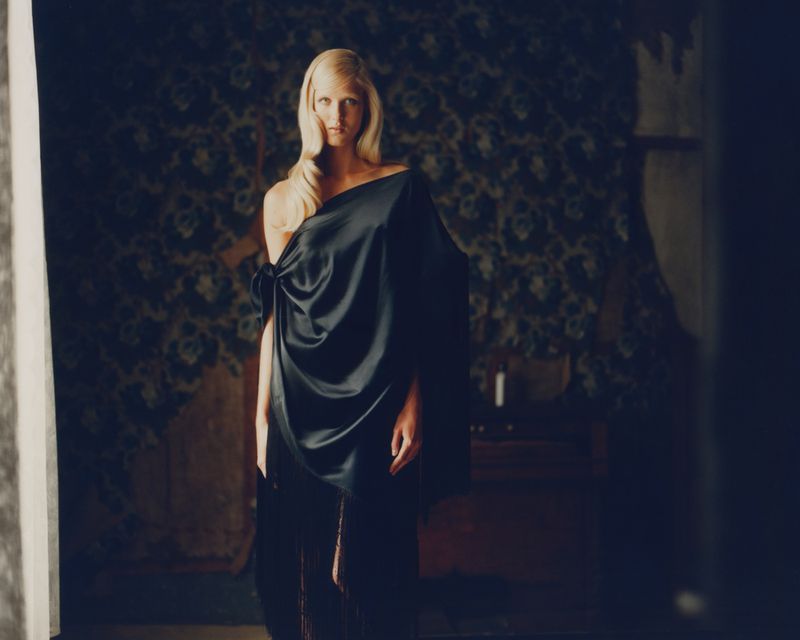 Adult woman with long blonde hair draped in a black dress posing in a moody interior with floral wallpaper, captured in a medium shot.