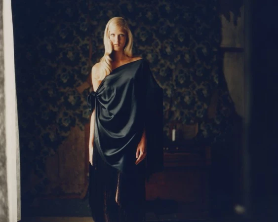 Adult woman with long blonde hair draped in a black dress posing in a moody interior with floral wallpaper, captured in a medium shot.