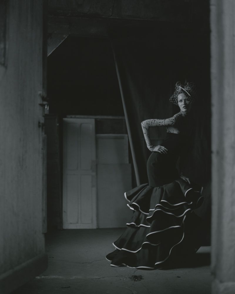 Elegant woman in long black dress with white ruffles poses in dimly lit hallway with white doors in the background in a vintage style.