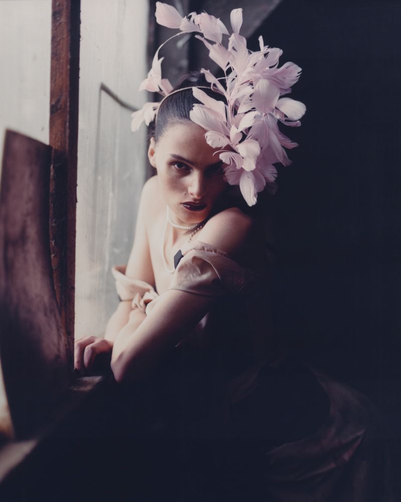 A woman wearing a headpiece with feathers looking at the camera. She is next to a window in a dark, moody atmosphere.