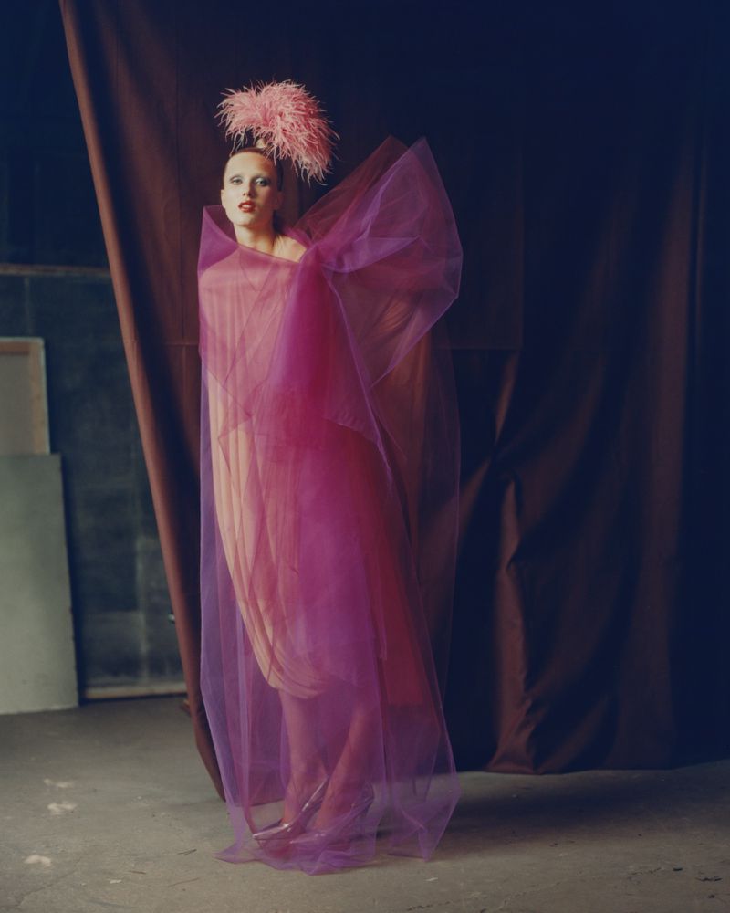 Elegant woman in a sheer magenta gown and feather headdress strikes a pose in an indoor studio with a draped brown backdrop.