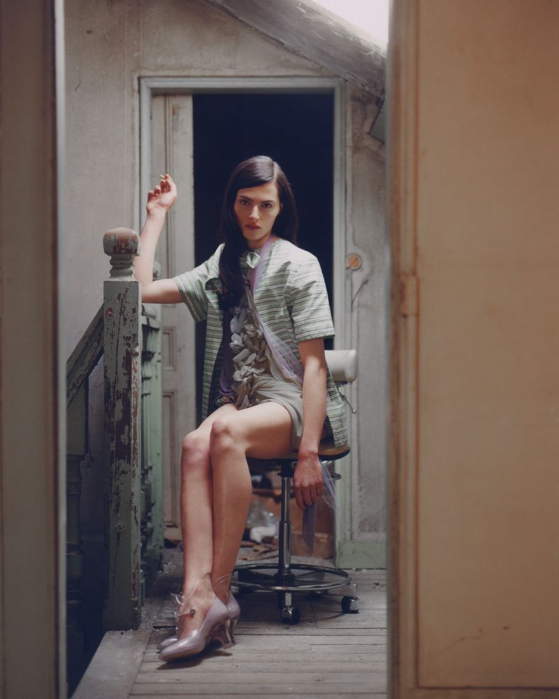 A female model poses on a rolling chair in a weathered doorway, wearing a green striped shirt, light shorts, and high heels in a worn interior.