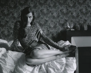 Model in leopard print top and belted skirt seated on rumpled white bed sheets against patterned floral wallpaper