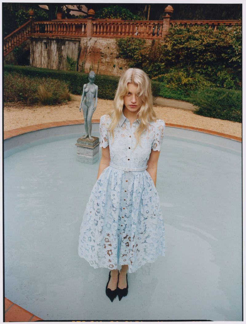 Model with blonde hair wearing a light blue lace dress standing inside a shallow outdoor fountain pool.