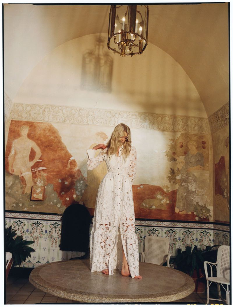 Model in a white lace maxi dress standing on a round stone platform inside a historic room with frescoed walls.