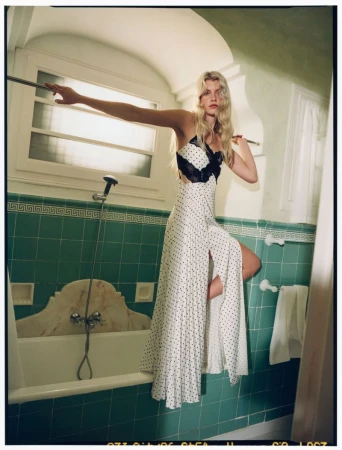 A woman poses in a polka dot dress in a vintage-style bathroom with green tiles and a white bathtub, bathed in soft, natural light.