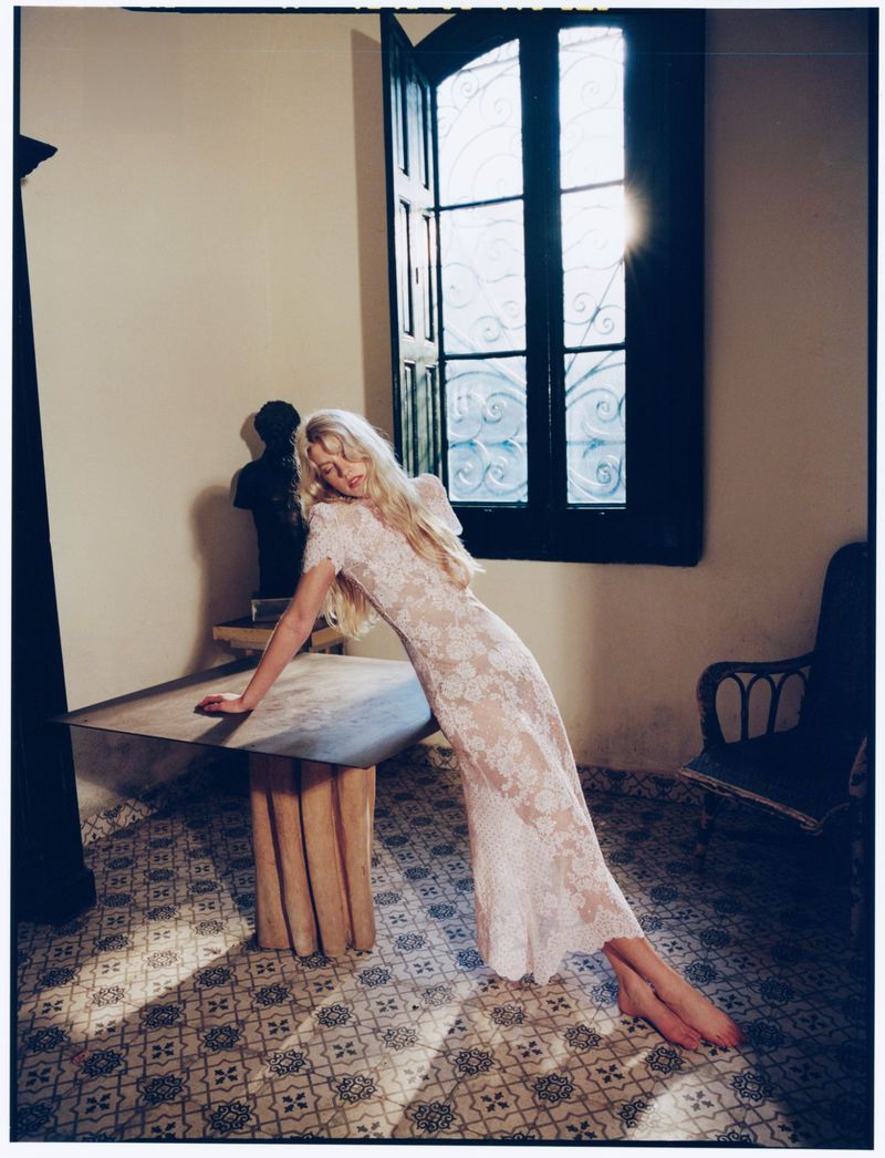 Model with long blonde hair wearing a lace dress leaning against a wooden table indoors with patterned tile flooring.