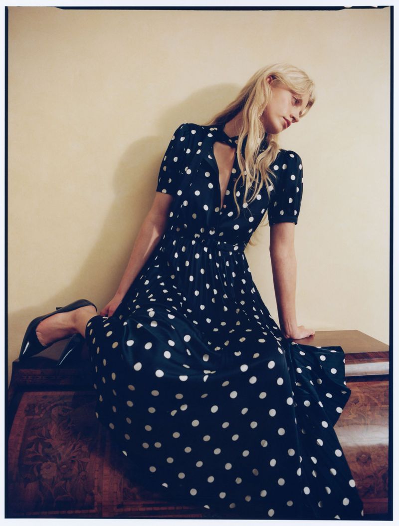Blonde model in a black polka dot dress seated on a wooden surface for Self-Portrait
