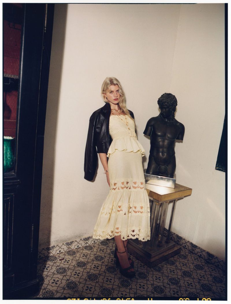 Model in a pale yellow strapless dress and black leather jacket posing next to a dark classical bust sculpture.