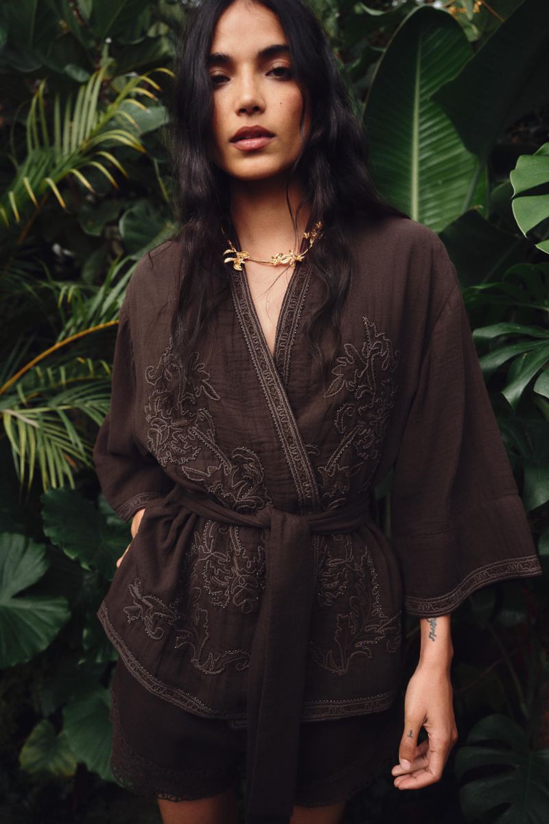 Model wearing an embroidered brown wrap jacket standing amidst lush tropical foliage for Zara