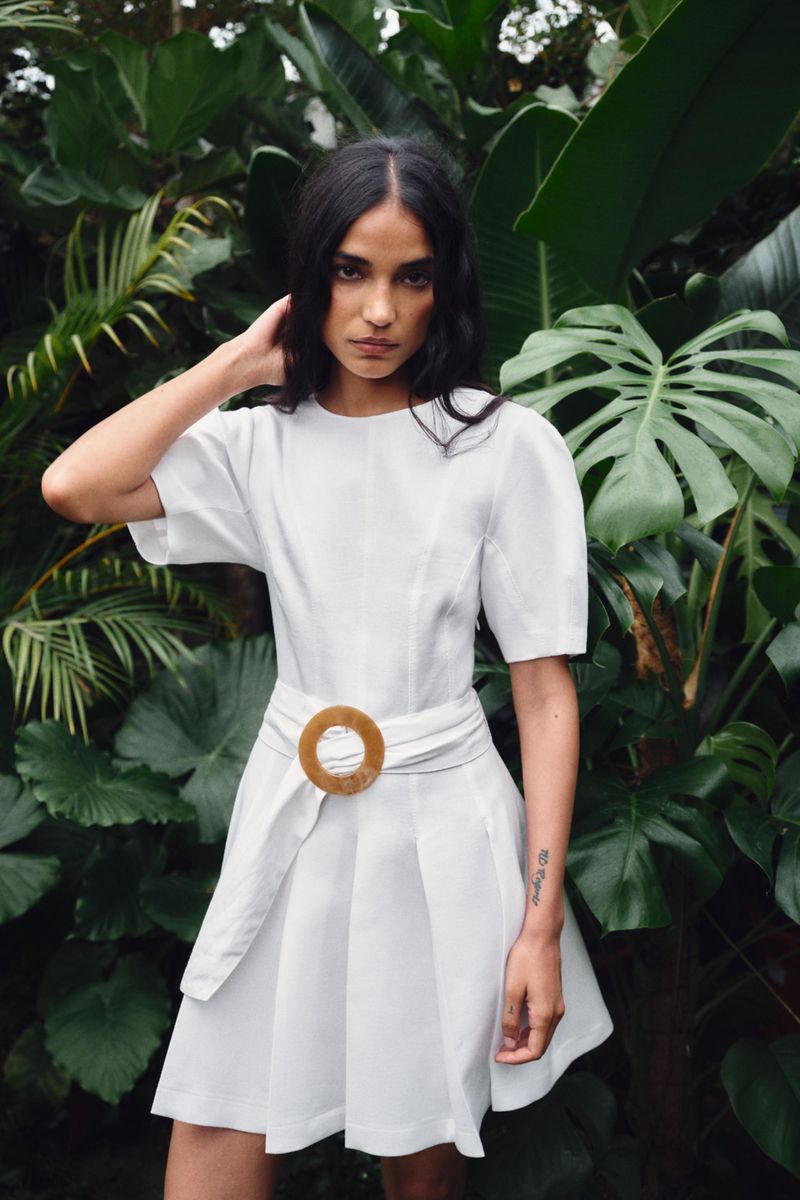 Model in white dress with belted waist standing amidst lush green tropical foliage