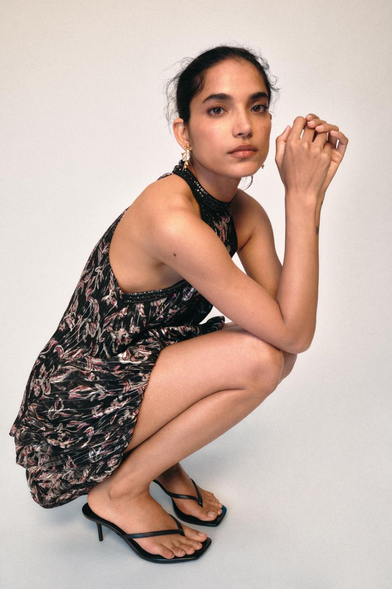 A young woman with dark hair poses in a studio wearing a short floral dress and black sandals while crouching against a plain white backdrop.